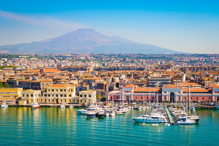 Port of Catania with turquoise blue sea, ocre and yellow buildings with terracotta roofs and Mount Etna behind